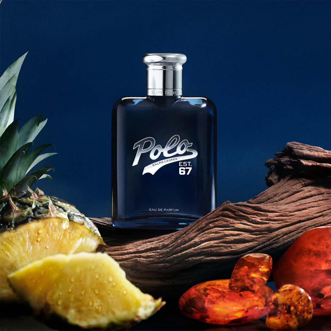 Polo 67 Eau de Parfum by Ralph Lauren bottle displayed with pineapple, amber resin, and driftwood on a dark blue background.